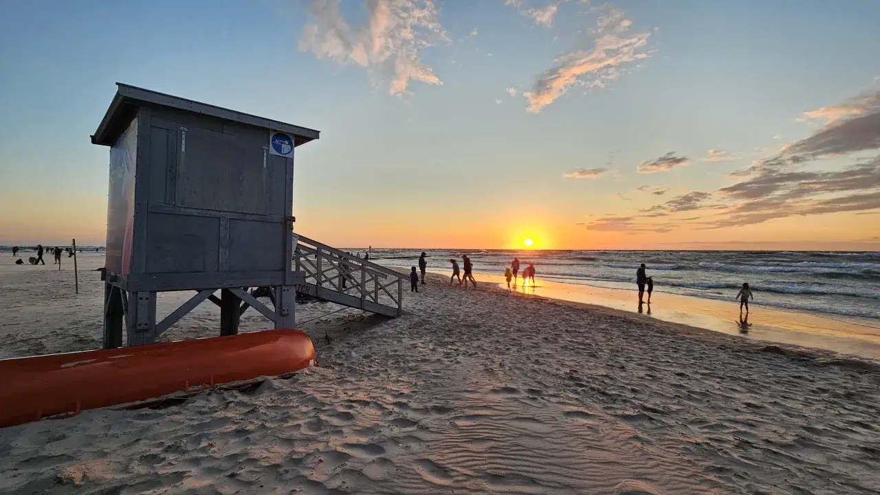 Zachód słońca nad morzem, ludzie spacerują po plaży, ratowniczy domek i pomarańczowa łódź. Idealne miejsce, co warto zobaczyć nad morzem.