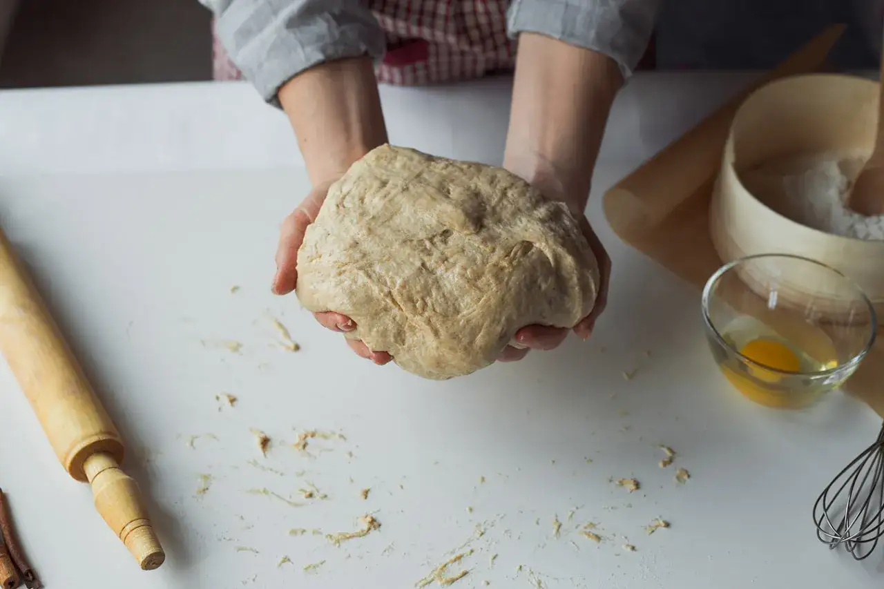 Ręce trzymające surowe ciasto, gotowe do dalszej obróbki. W tle wałek, mąka i jajko – wszystko, co potrzebne, by dowiedzieć się, jak mrozić ciasto.