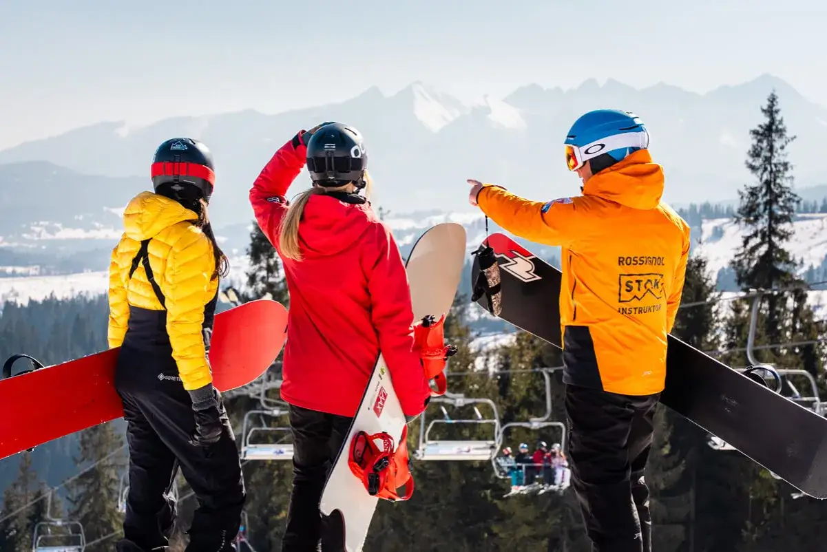 Instruktor w pomarańczowej kurtce pokazuje kierunek, odpowiadając na pytanie: ile trwa nauka jazdy na nartach. Trzy osoby ze snowboardami na tle gór.