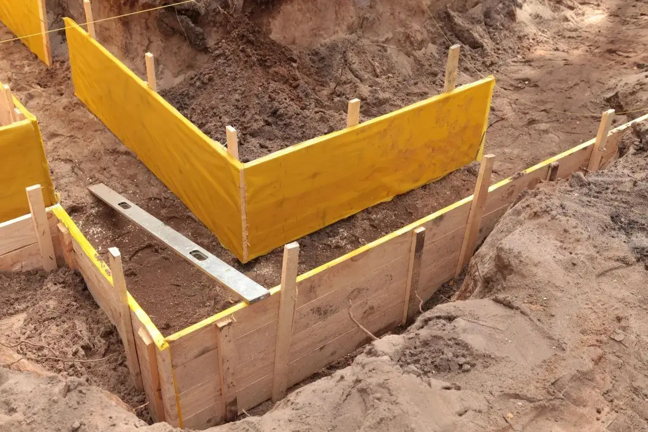 Wykonano wykop pod fundament, widać drewniane szalunki i żółtą folię. Poziomica leży na ziemi.