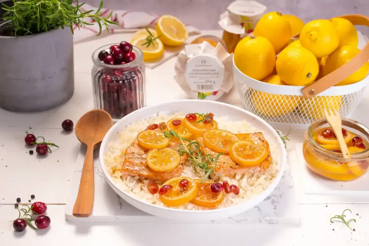 Pyszny łosoś na ryżu, udekorowany plasterkami cytryny i żurawiną. Obok koszyk z cytrynami i słoiczek z marynowanymi cytrynami.