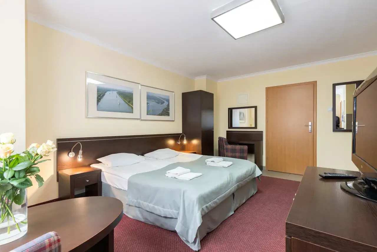 Przytulny pokój w resort spa plaza: łóżko, szafka nocna z lampką, szafa, biurko, lustro, drzwi i telewizor.