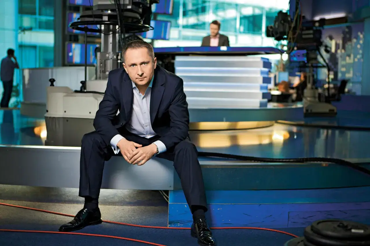 Mężczyzna w garniturze siedzi w studiu telewizyjnym. Czy to dlatego znany dziennikarz zmienił nazwisko przed rozpoczęciem kariery?