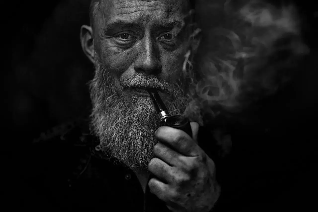 Intensywny portret mężczyzny z siwą brodą, palącego fajkę. Dym tworzy mistyczną aurę, podkreślając r&oacute;żne rodzaje portret&oacute;w w fotografii.