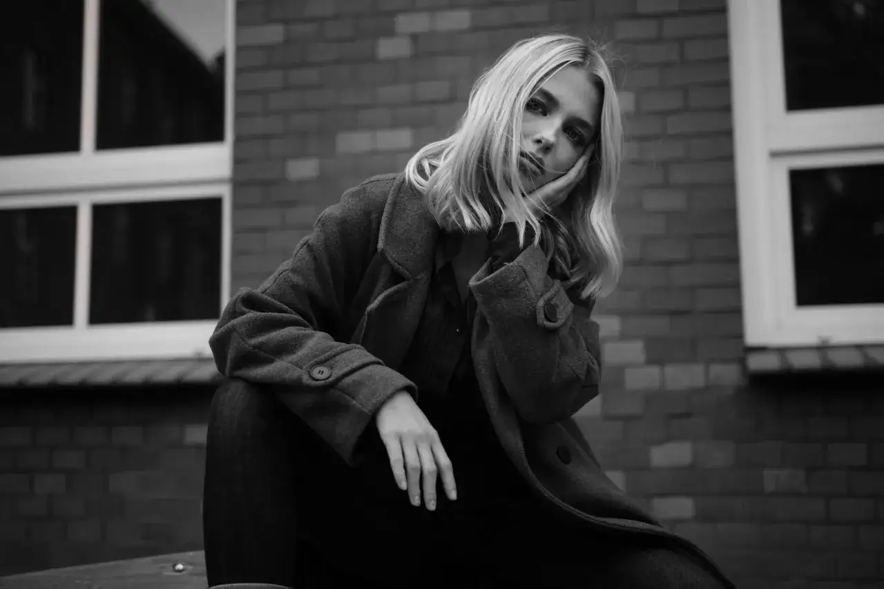 Smutna blondynka w płaszczu opiera głowę na dłoni. Ten kadr uchwycił jej melancholijny nastrój na tle ceglanej ściany i okien.