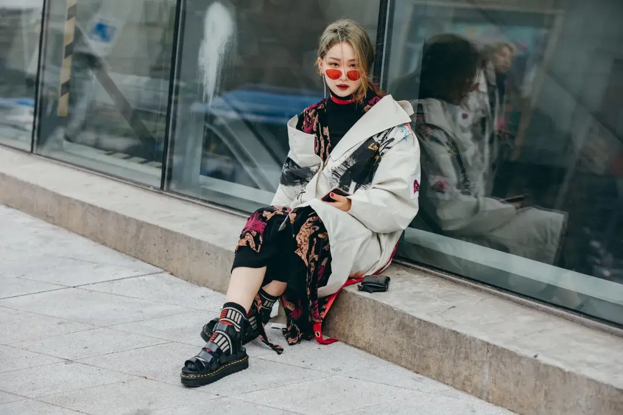 Modna dziewczyna w okularach przeciwsłonecznych i wzorzystej sukience, z czarnymi sandałami i kolorowymi skarpetami, siedzi na betonowej ławce.