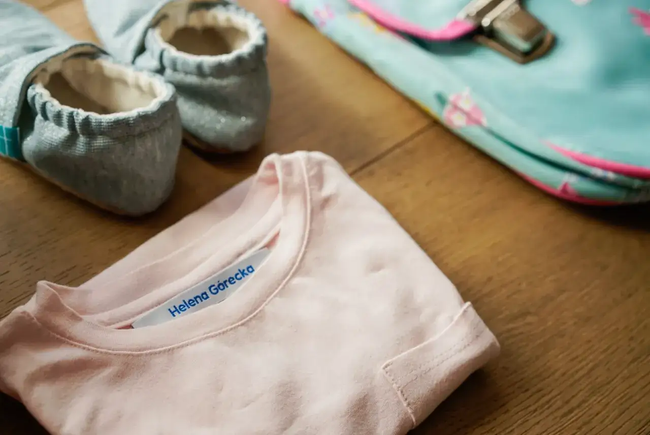 Jak podpisać ubrania do żłobka? Różowa koszulka z imieniem Helena Górecka, kapcie i torba na drewnianym tle.