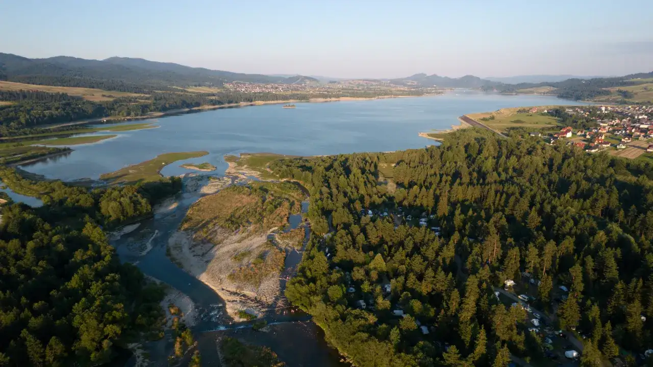 Widok z lotu ptaka na jezioro i las, gdzie rozbity jest camping łęg frydman. Widać namioty, kampery i samochody wśród drzew.