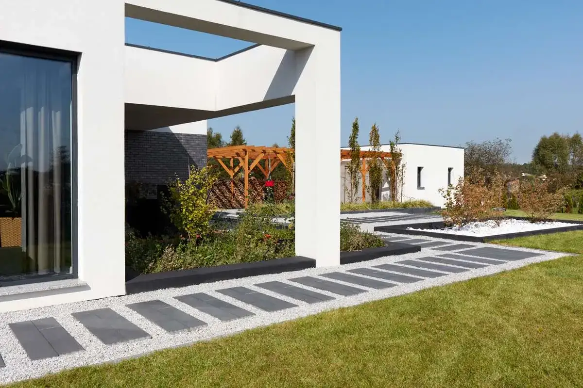 Nowoczesny dom z podjazdem bez ogrodzenia, wyłożonym kamieniami i płytami. Wokół zieleń i drewniana pergola.