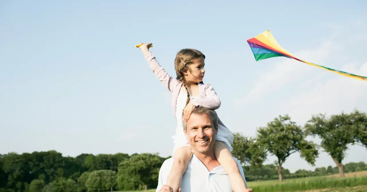 Ojciec niesie c&oacute;rkę na barana, a ona puszcza tęczowego latawca. Wspaniała zabawa na świeżym powietrzu, jak zrobić latawiec to świetny pomysł na rodzinne popołudnie.