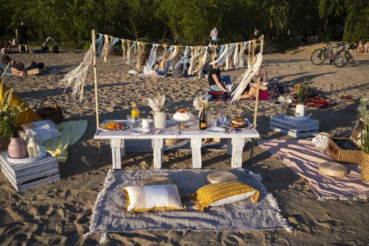 St&oacute;ł z ciastem, owocami i napojami, poduszki i koce na piasku. Idealne miejsce, by zobaczyć, co zabrać na piknik.