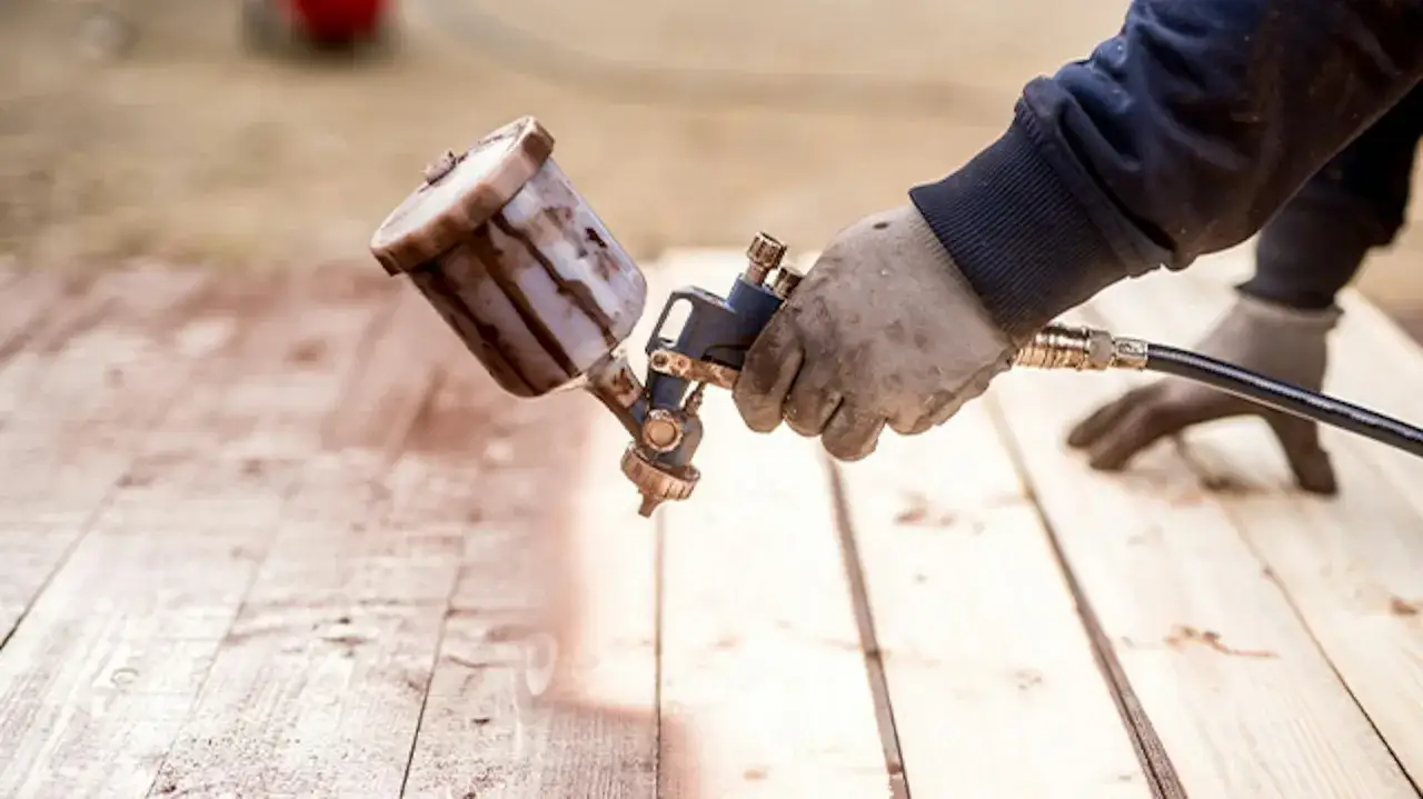 Ręka w rękawicy trzyma pistolet natryskowy, aplikując brązowy impregnat do drewna na zewnątrz.