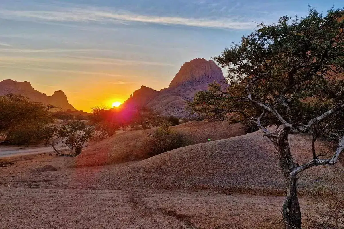 Zach&oacute;d słońca nad Spitzkoppe w Namibii, z charakterystycznymi skalnymi formacjami i drzewami akacjowymi.