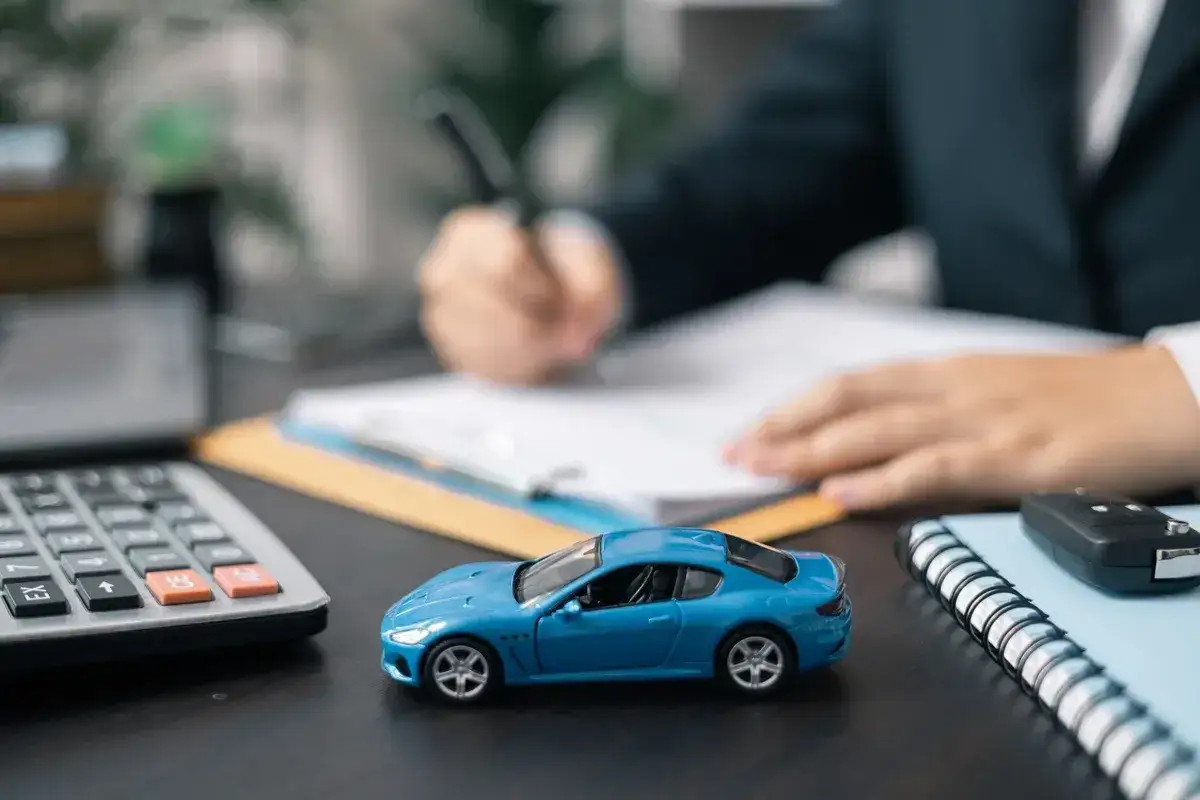 Niebieski samochodzik, kalkulator i kluczyki na biurku symbolizują leasing finansowy. Dłoń pisze umowę.