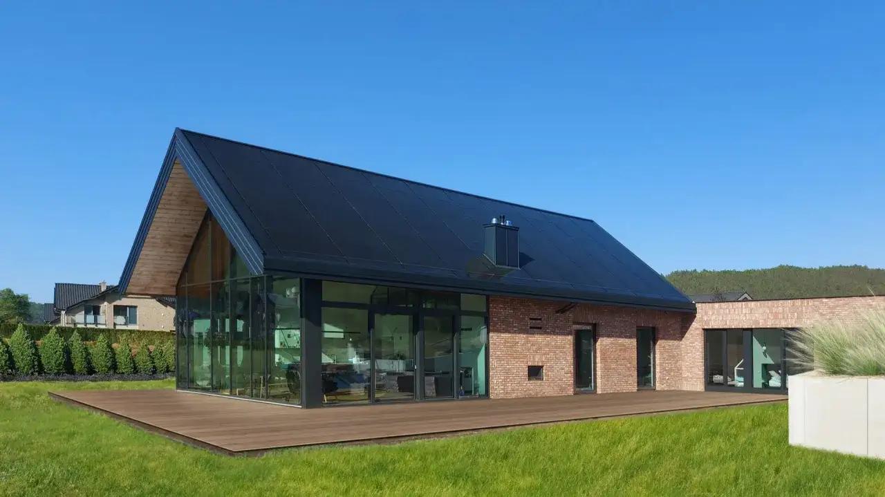 Nowoczesny dom z cegły i szkła, z imponującym dachem z paneli fotowoltaicznych, otoczony zielonym trawnikiem i drewnianym tarasem.