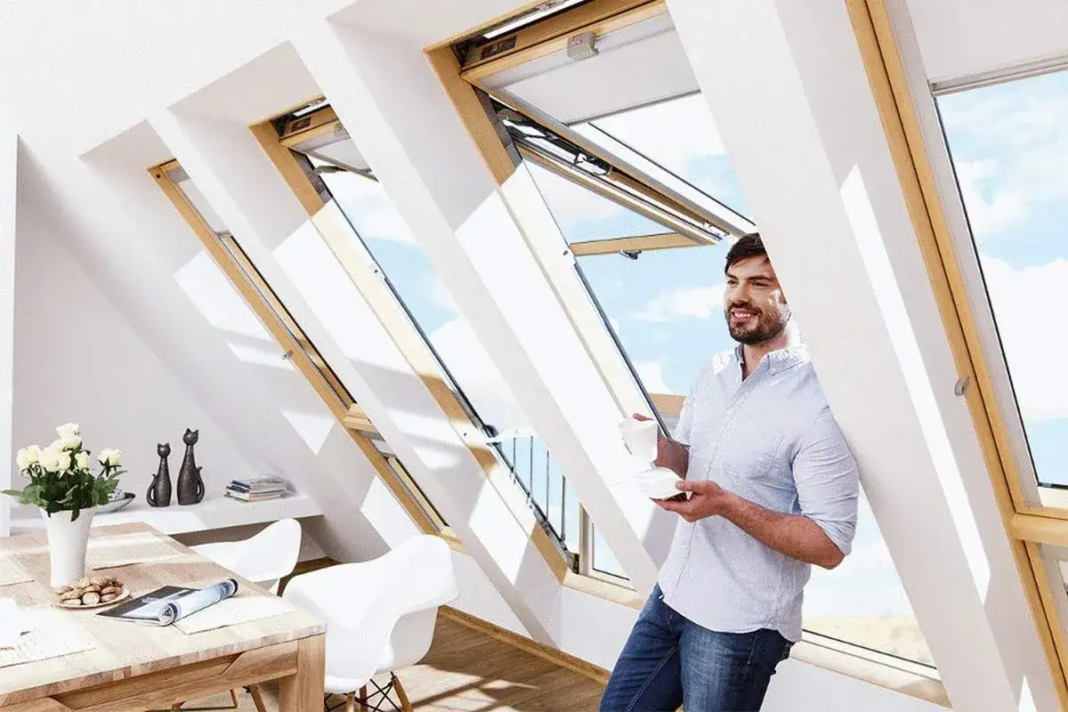 Mężczyzna z kawą opiera się o ścianę, ciesząc się widokiem przez otwarte okno dachowe balkonowe.