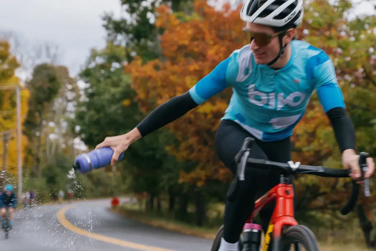 Kolarz w niebieskiej koszulce i czarnych getrach, z kaskiem i okularami, pokazuje, jak się ubrać na rower, chłodząc się wodą z bidonu.