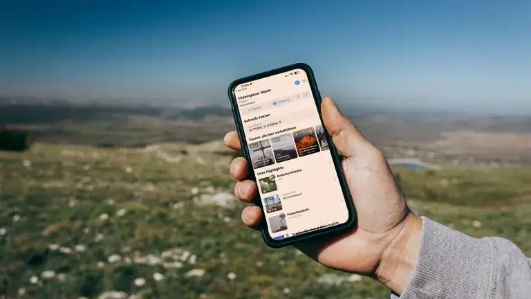 Person benutzt Wander-App auf Smartphone mit Bergpanorama im Hintergrund