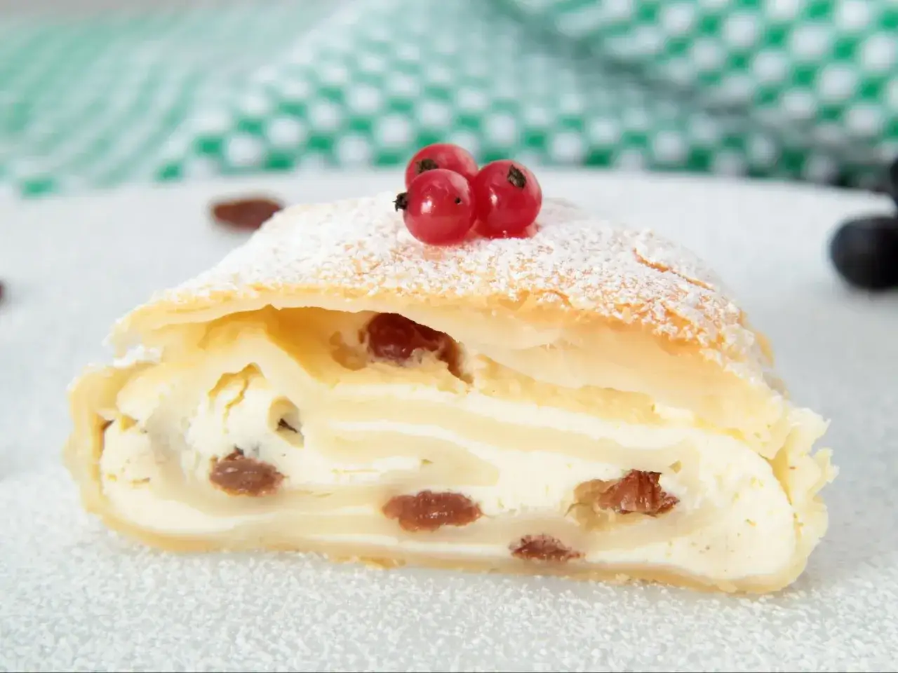 Pyszna rolada serowa z rodzynkami, posypana cukrem pudrem i udekorowana czerwonymi porzeczkami.