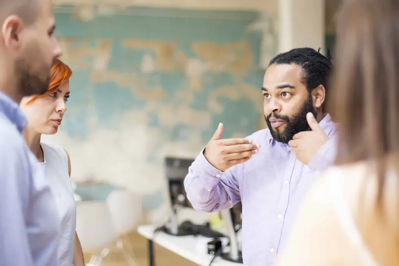 Mężczyzna z dredami i brodą gestykuluje, omawiając ciekawe tematy do rozmowy z kobietą o rudych włosach i mężczyzną w jasnej koszuli.