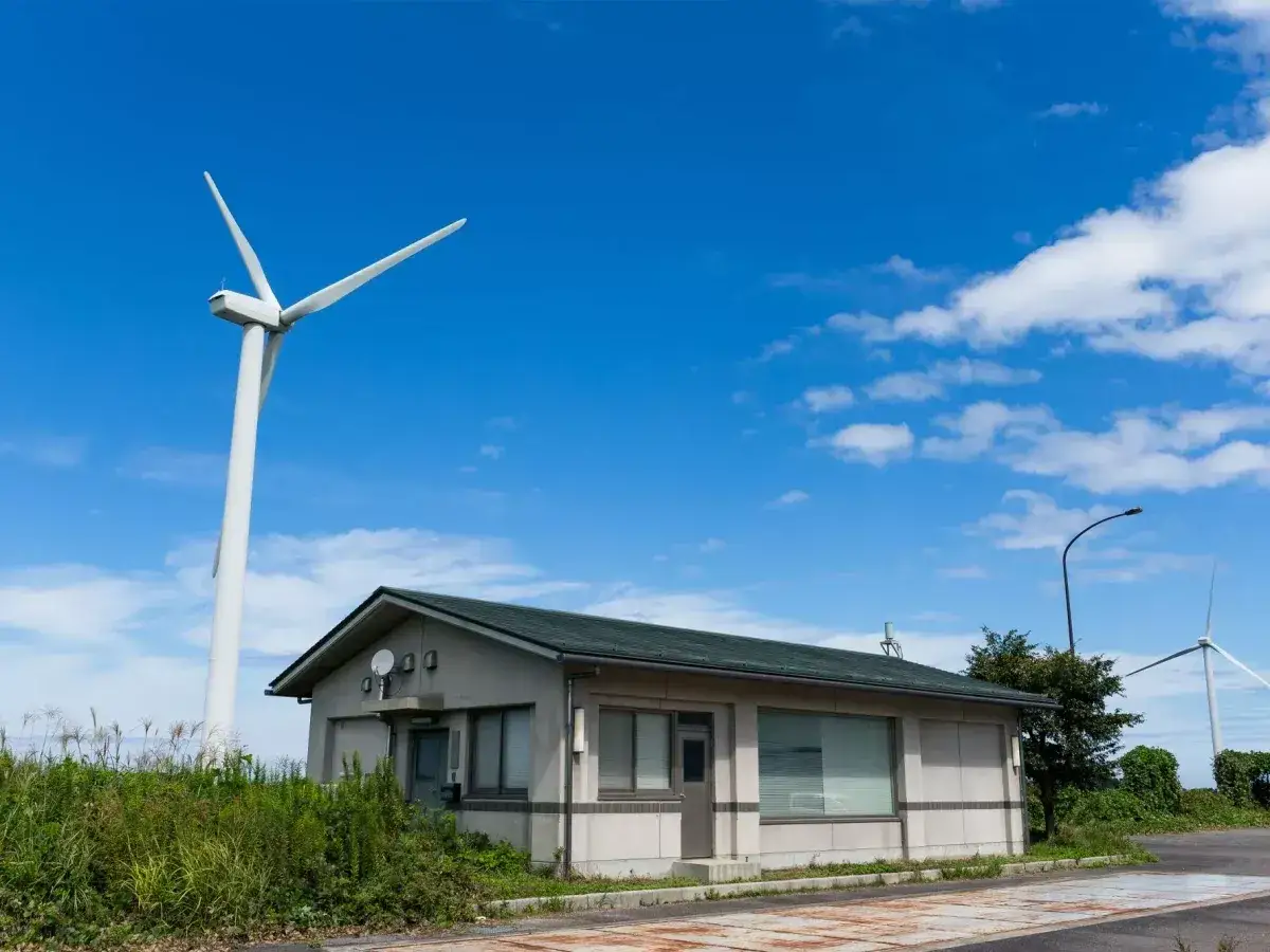 Biała turbina wiatrowa bez pozwolenia g&oacute;ruje nad małym budynkiem. W tle błękitne niebo z chmurami.