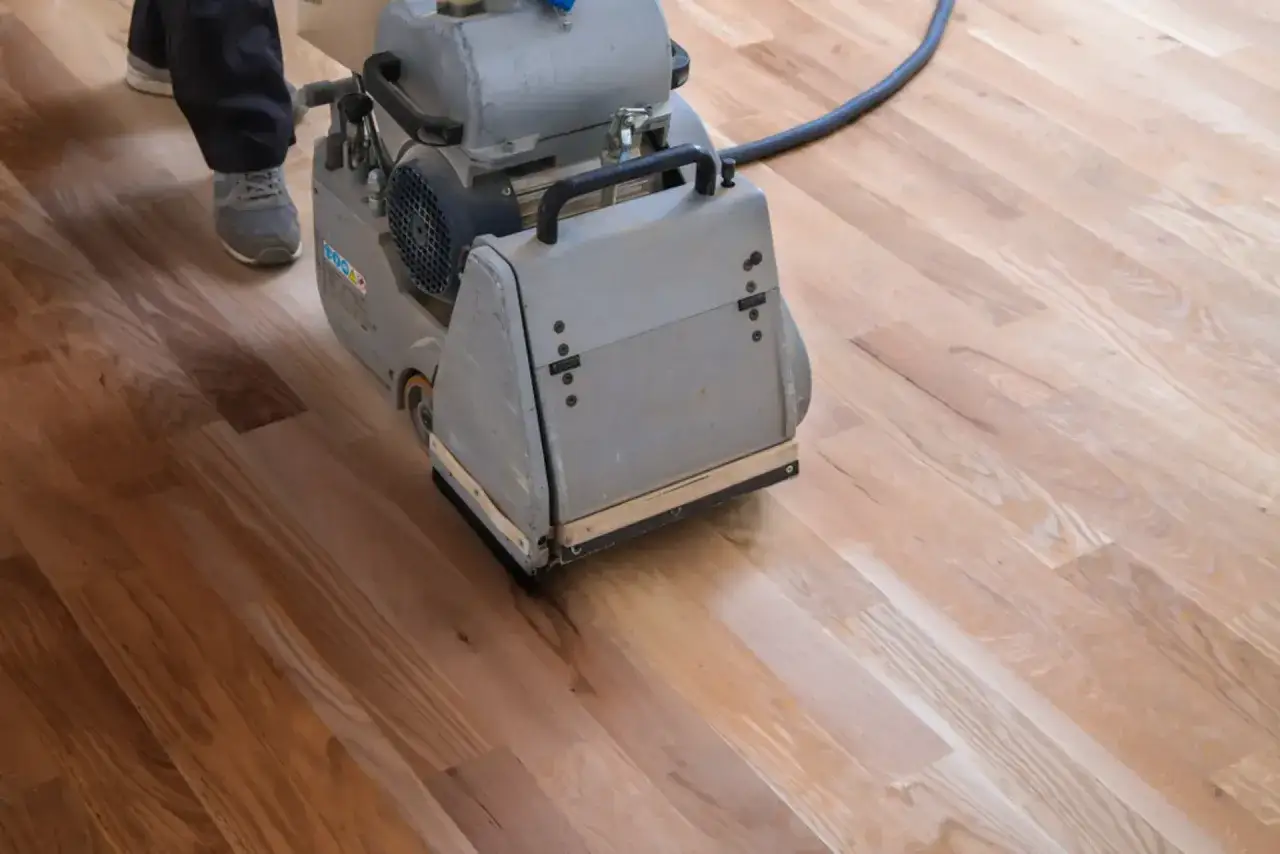 Maszyna do cyklinowania przygotowuje parkiet do lakierowania. Widać fragmenty drewnianej podłogi i nogi osoby obsługującej sprzęt.