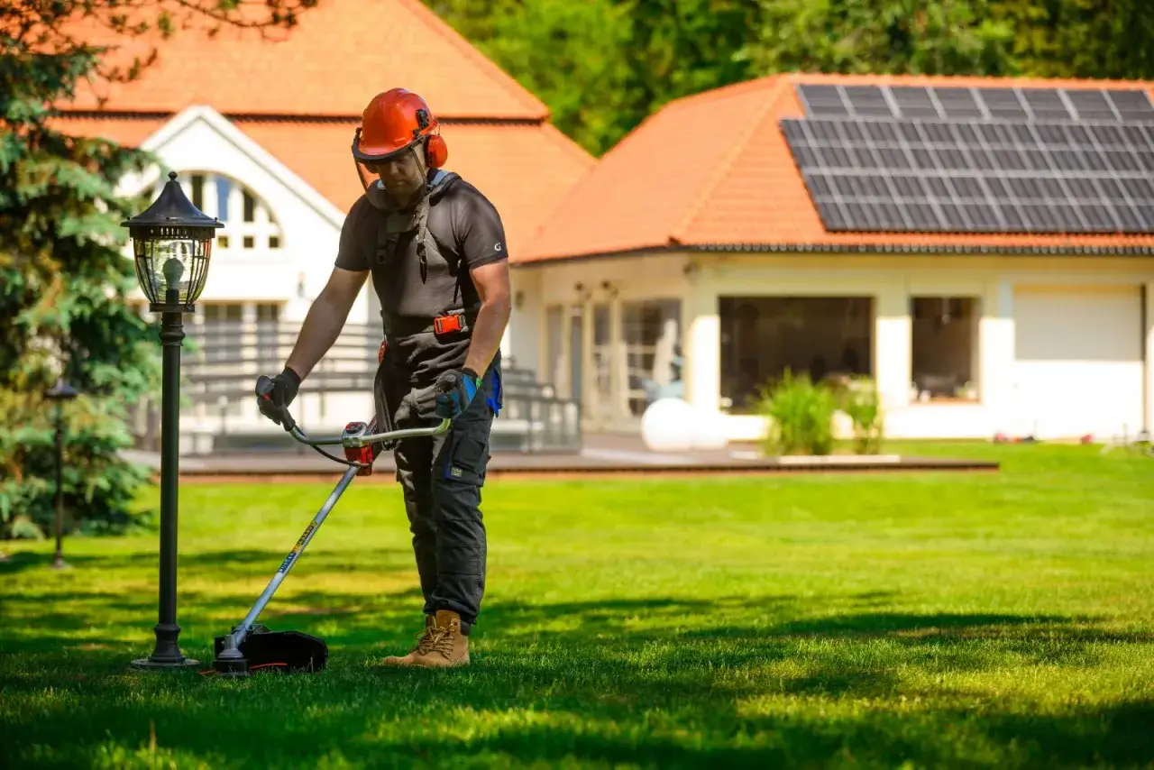Mężczyzna w kasku i słuchawkach pokazuje, jak kosic kosa spalinowa, dbając o piękny trawnik przed domem z panelami słonecznymi.