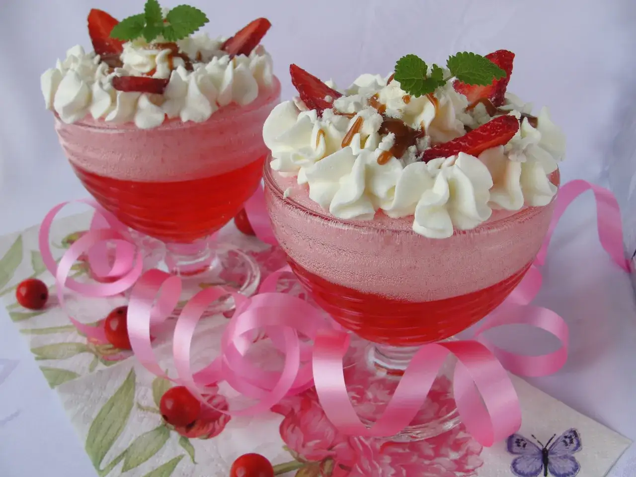 Jak zrobić piankę z galaretki? Dwa pucharki z deserem truskawkowym, bitą śmietaną, owocami i polewą.