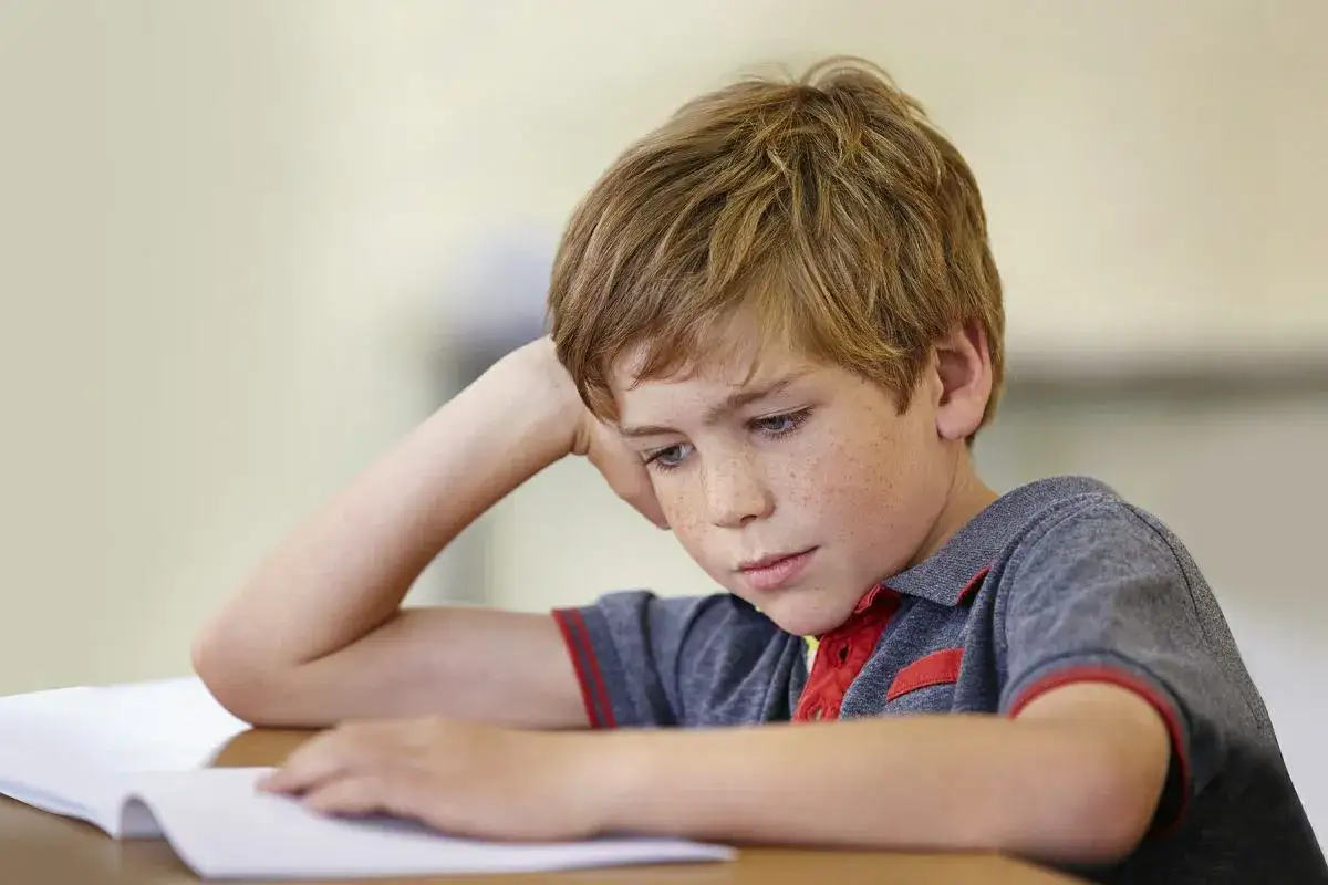 Chłopiec z piegami pochyla się nad książką, zamyślony. Czy dysleksja to choroba, która utrudnia mu naukę?