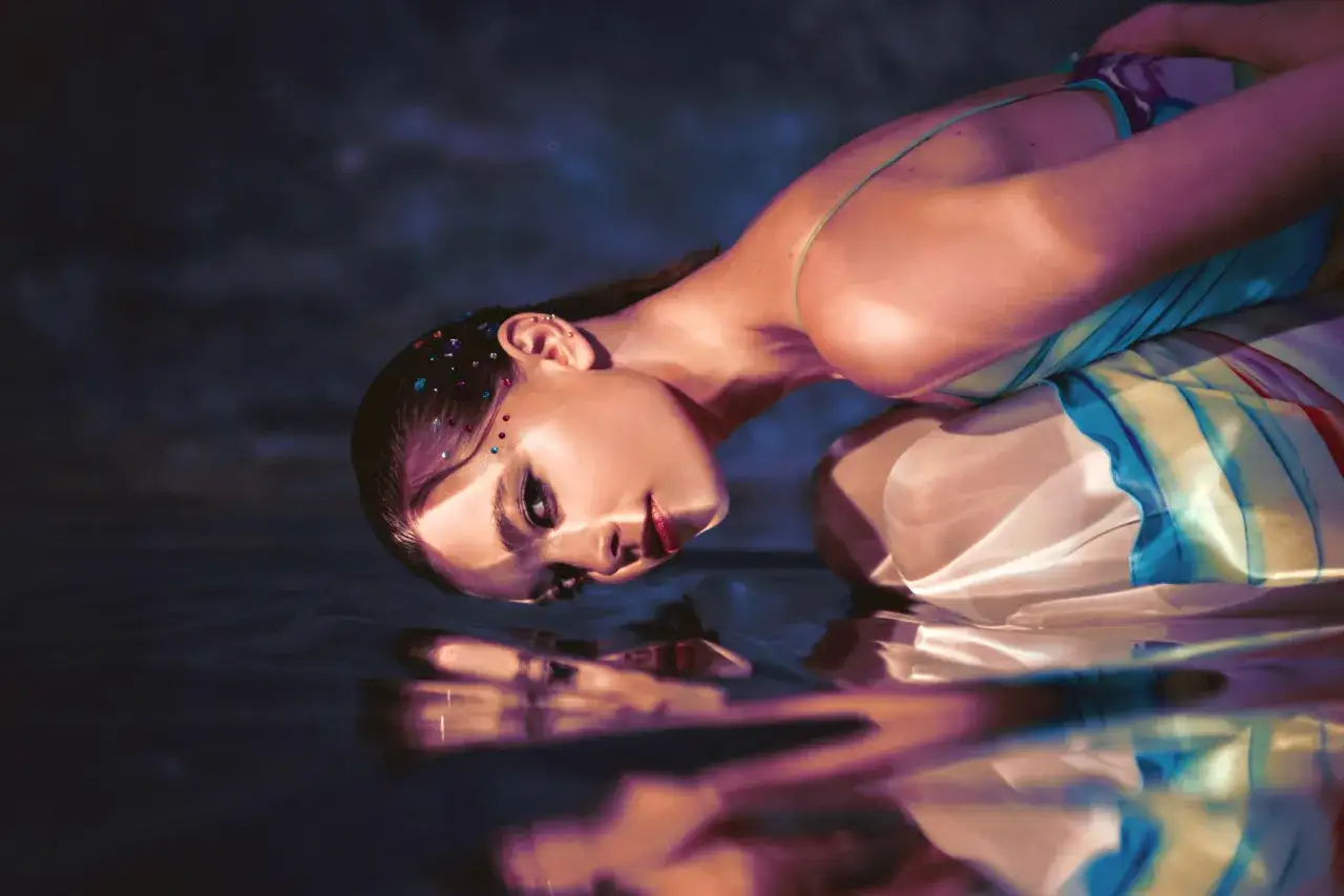 Artystyczna fotografia beauty. Modelka z błyszczącymi kryształkami na twarzy, w kolorowej sukni, zanurzona w wodzie.