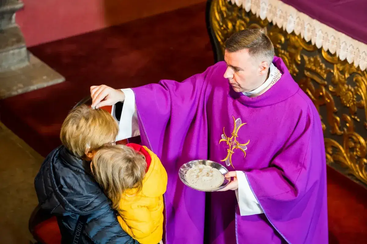 Ksiądz w fioletowej dalmatyce posypuje popiołem głowy dzieci podczas mszy świętej w środę popielcową.