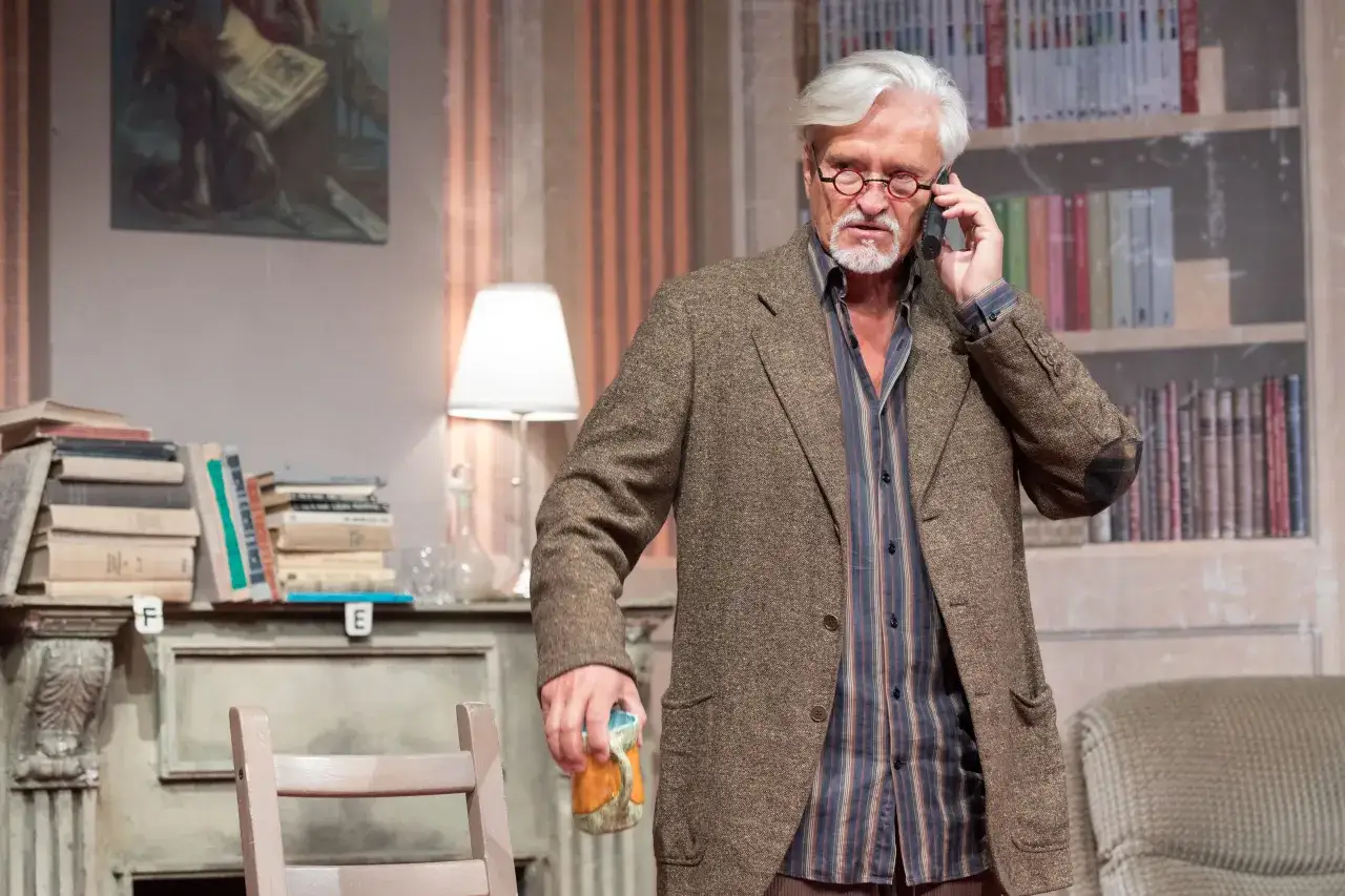 Starszy mężczyzna w tweedowej marynarce rozmawia przez telefon, trzymając kubek. Scenografia sugeruje edukację i spektakl.