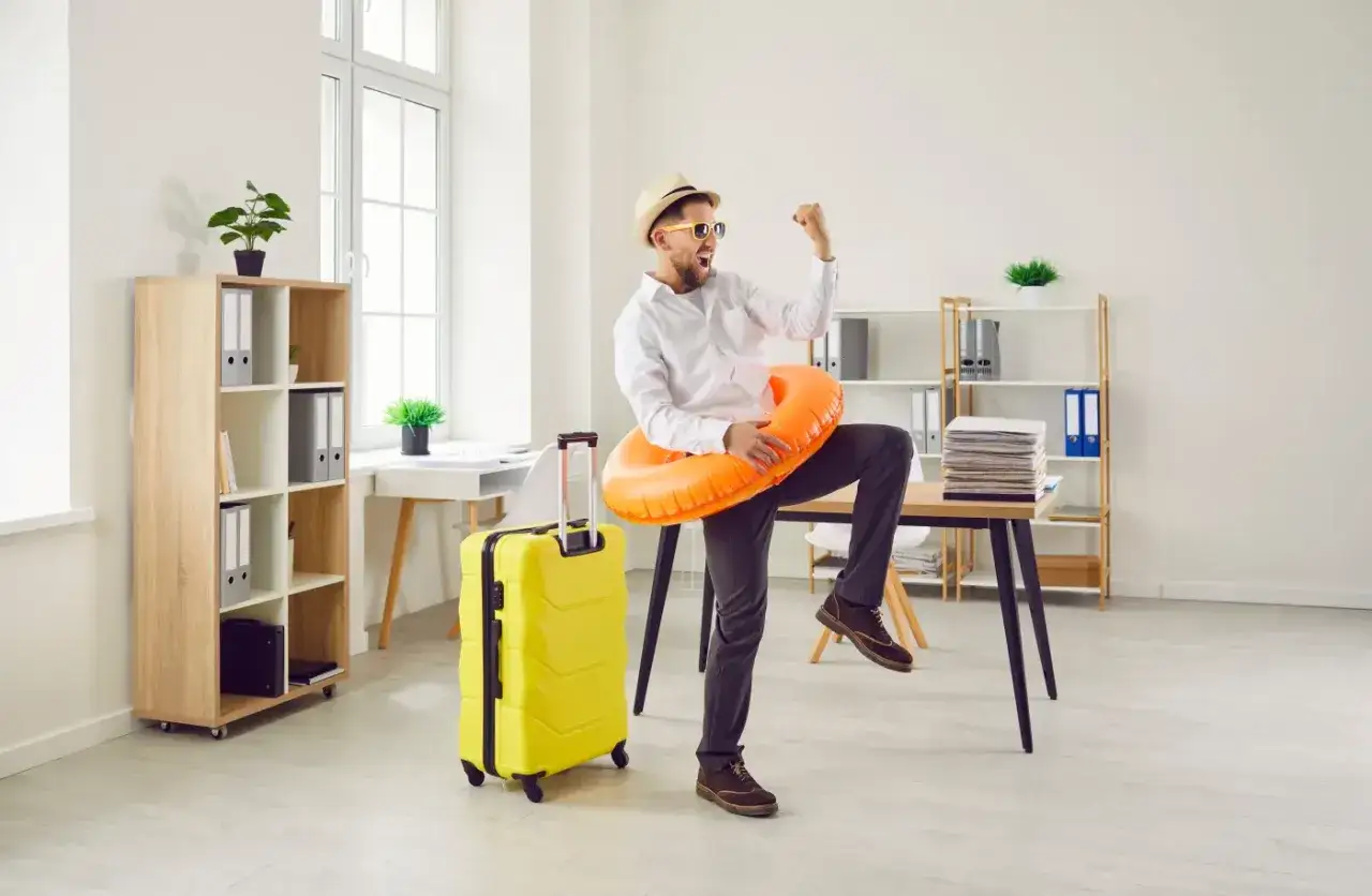 Radosny mężczyzna w kapeluszu i okularach przeciwsłonecznych, z kołem ratunkowym na szyi i walizką, świętuje koniec pracy. Czy urlop bezpłatny wlicza się do emerytury?