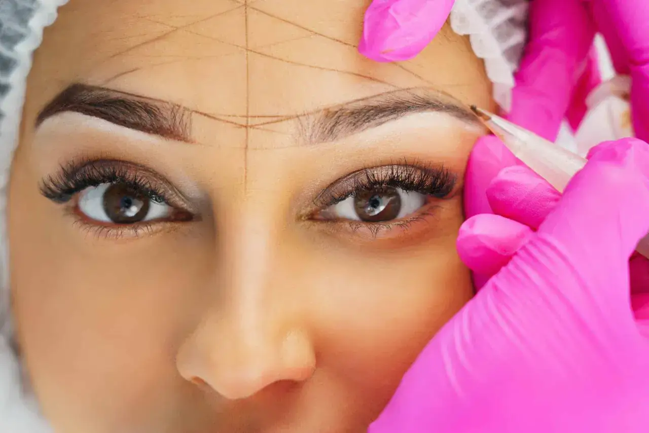 Nahaufnahme einer Frau, deren Augenbrauen mit Linien markiert werden, während eine Person in pinken Handschuhen mit einem Stift arbeitet.