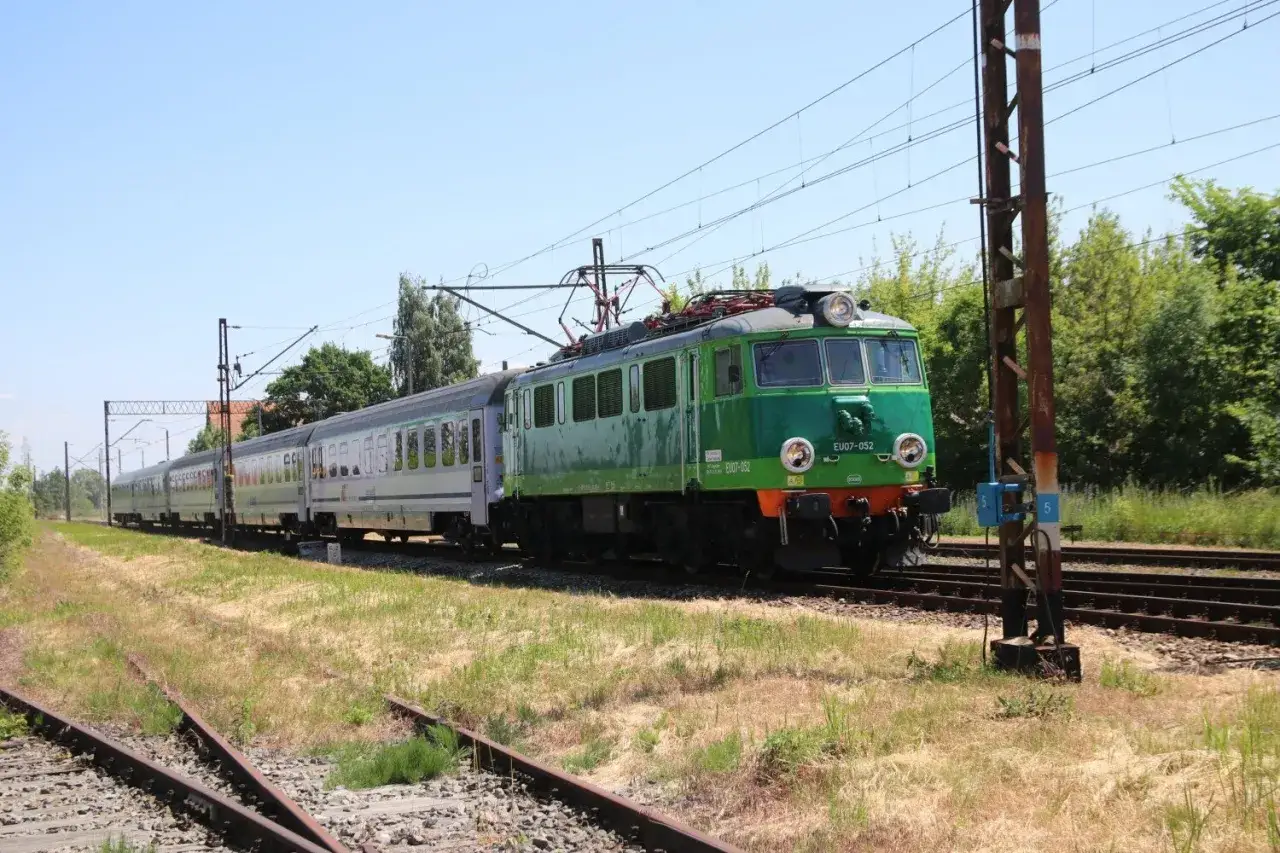 Zielony pociąg EU07-052 z wagonami mija tory w słoneczny dzień. To widok z wieluń trasa.
