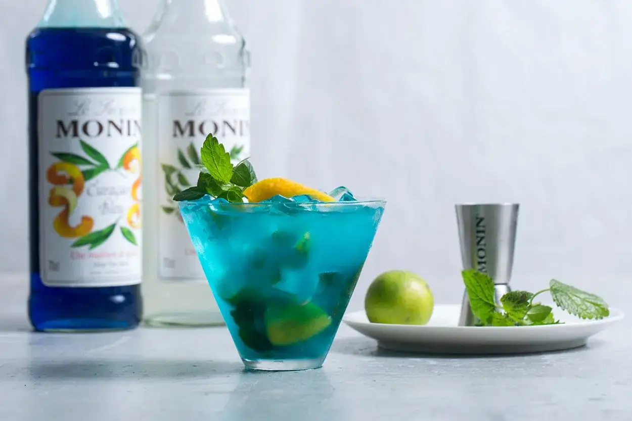 Orzeźwiający drink z syropem blue curacao smak, ozdobiony miętą i cytryną. Obok butelki syropu Monin.