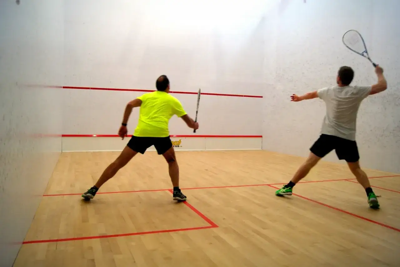 Dwóch mężczyzn gra w squasha na boisku z drewnianą podłogą i białymi ścianami.