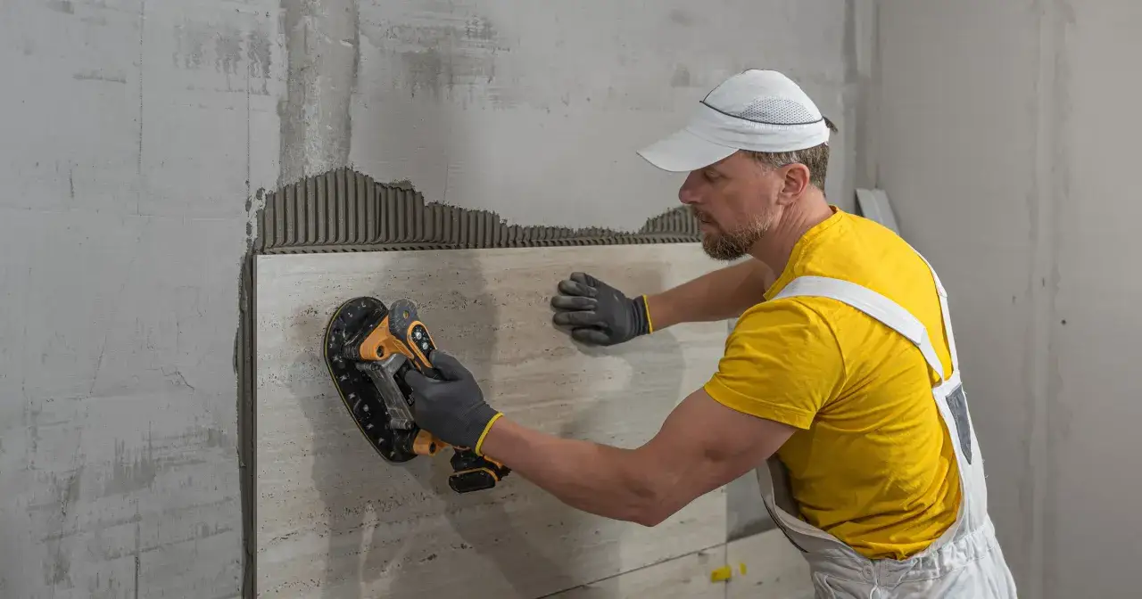 Fachowiec z narzędziem do układania płytek, w żółtej koszulce i białym ogrodniczkach, kładzie płytki na ścianie. To ulga na remont!