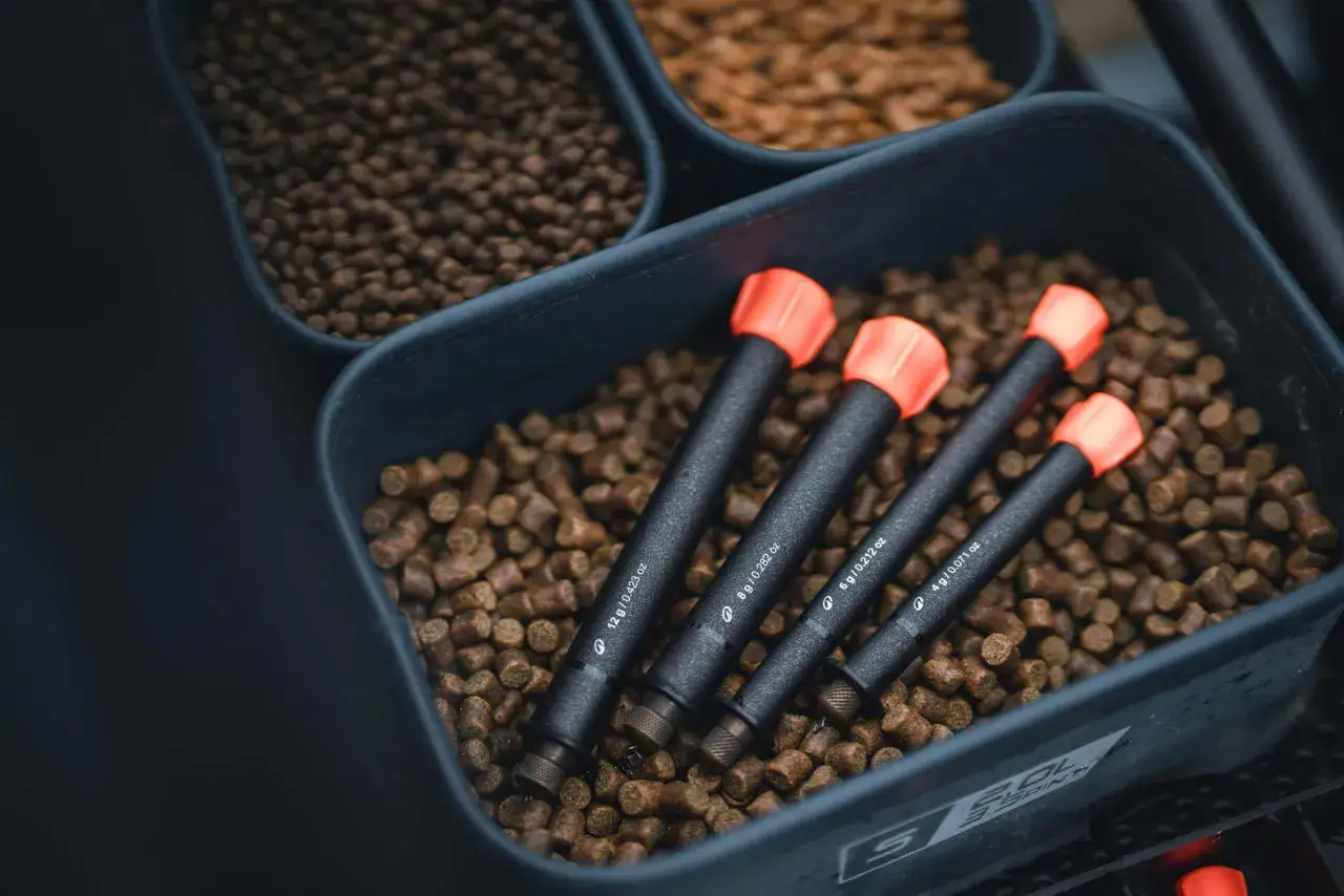 Zanęta halibut na jakie ryby? W pojemniku z pelletem leżą cztery sygnalizatory brań z pomarańczowymi końcówkami.