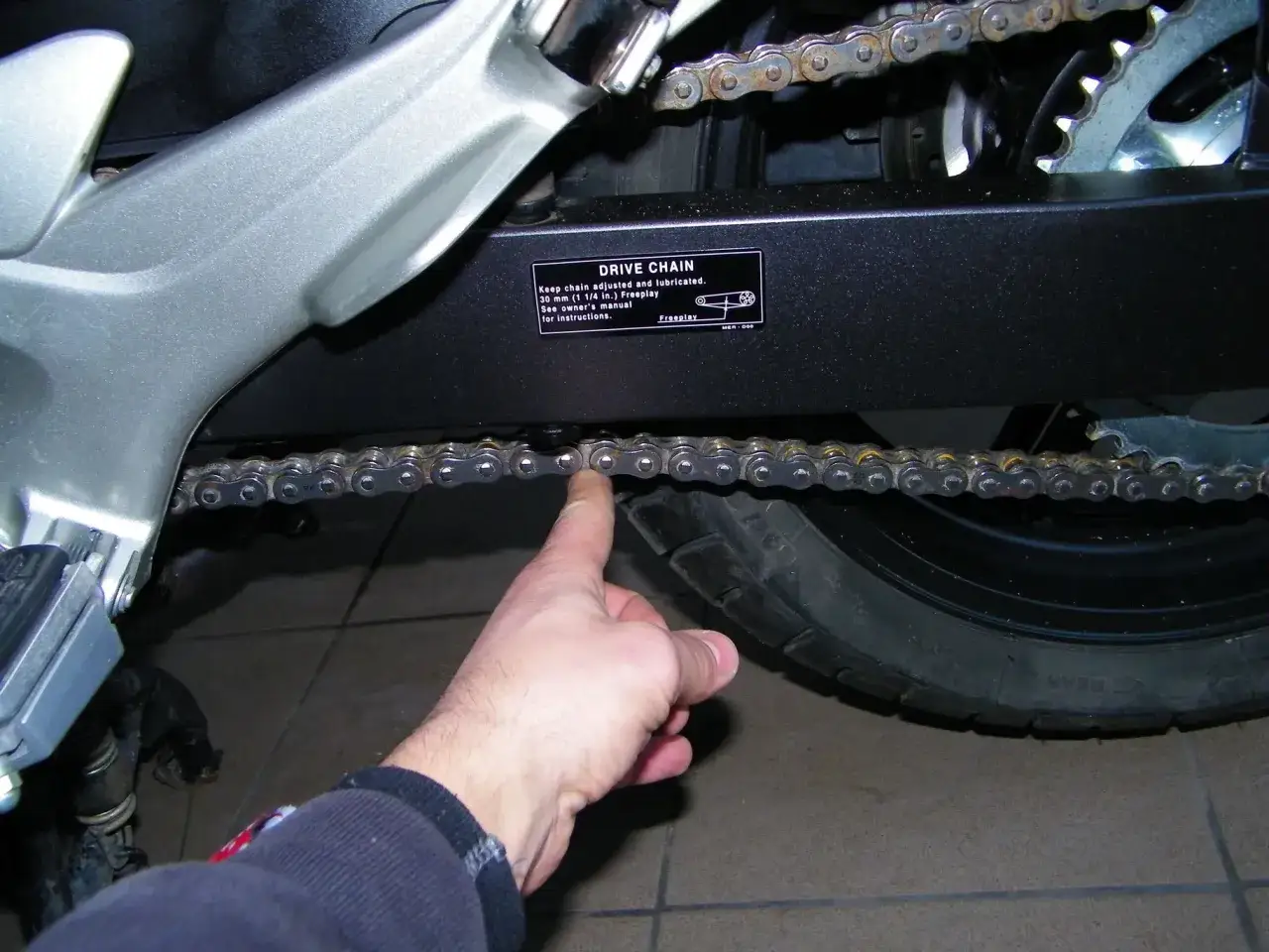 Dłoń wskazująca na luźny łańcuch motocyklowy przy wahaczu.