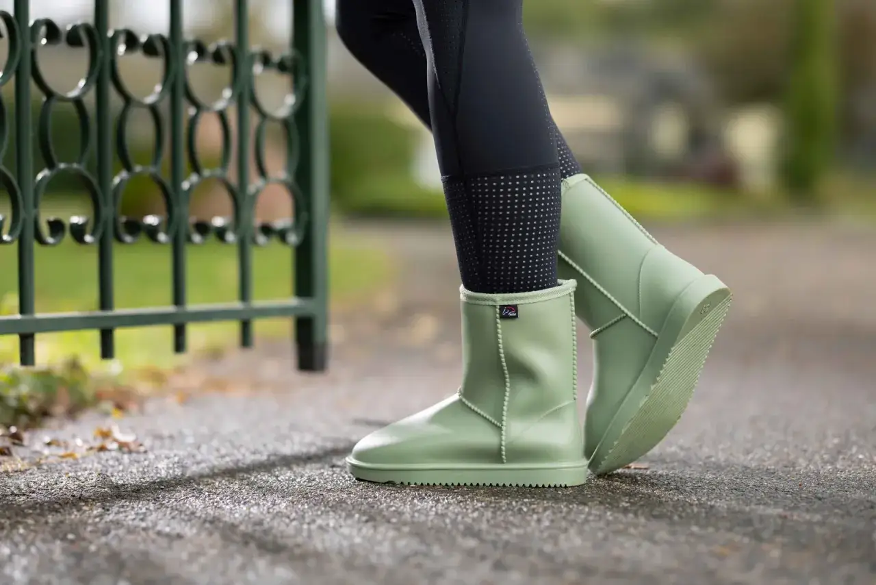 Jasnozielone, gumowe buty typu UGG na nogach w czarnych legginsach, obok zielonego ogrodzenia.