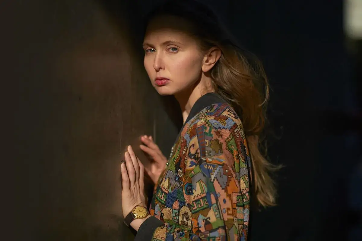 Justyna Wasilewska w kolorowej kurtce opiera się o ciemną ścianę, jej twarz oświetla cień.