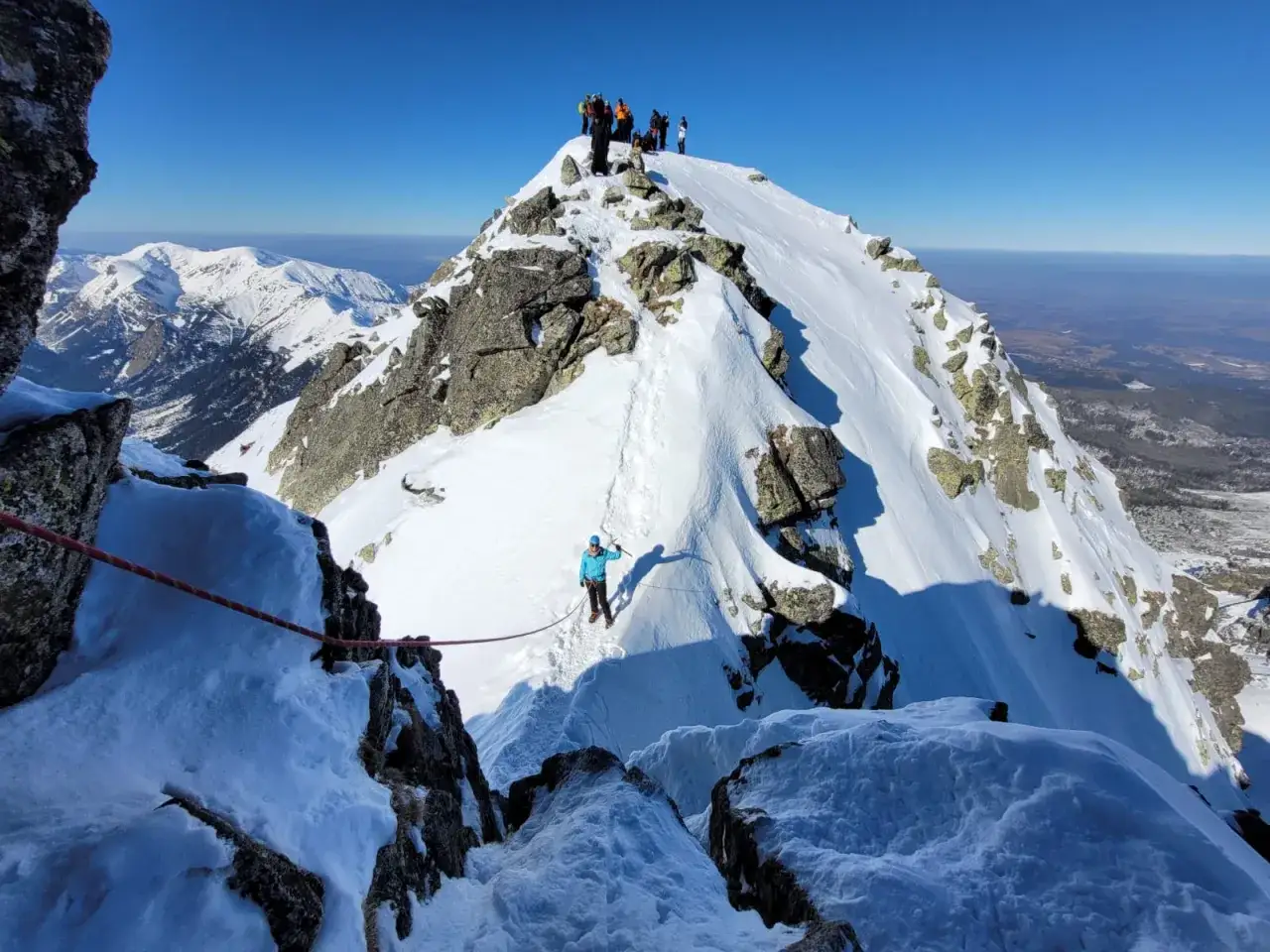 Grupa turystów na ośnieżonym szczycie. Czy szlak na Świnicę jest otwarty? Widać linę asekuracyjną i ślady na śniegu.