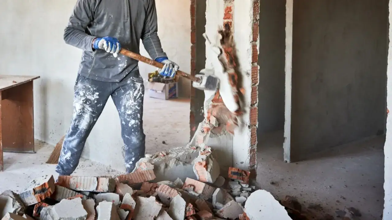 Mężczyzna w rękawicach i ubraniu w plamach farby burzy ścianę młotem. Czy spółdzielnia może zabronić remontu mieszkania?