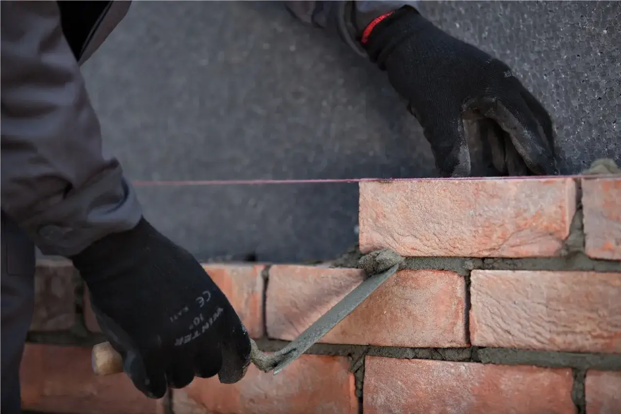 Ręce w czarnych rękawicach wykonują murowanie ścian działowych, układając cegły na zaprawie.