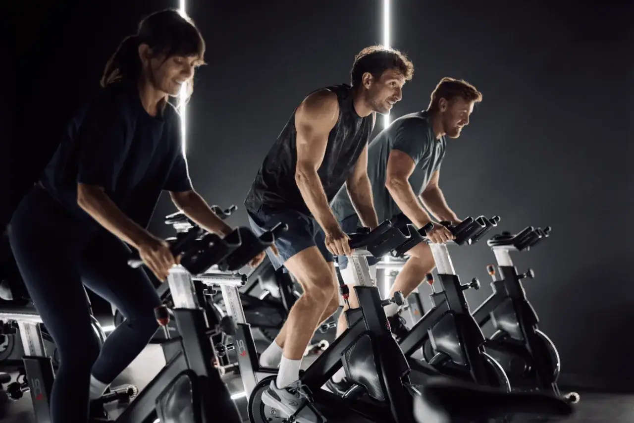 Trzy osoby ćwiczą na rowerach stacjonarnych w ciemnym pomieszczeniu. Widać ich skupienie i wysiłek, co sugeruje pozytywne indoor cycling efekty.