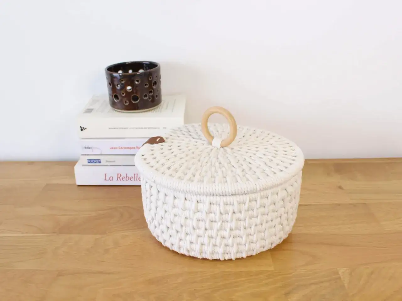 Biały koszyk z makramy bez szydełka, z drewnianą rączką, stoi obok stosu książek i ozdobnego świecznika.