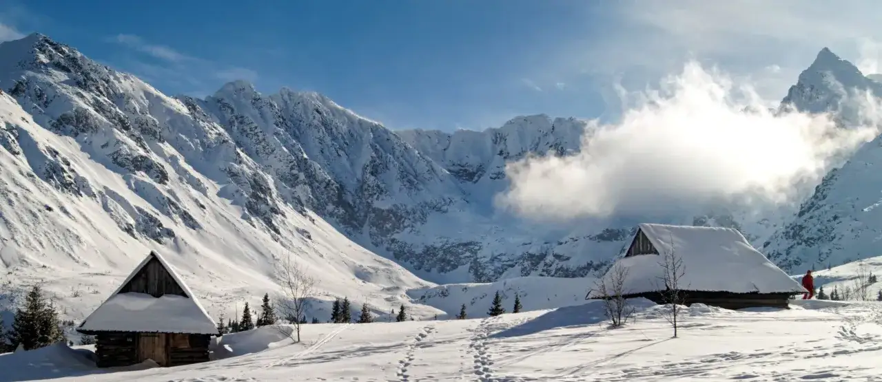 Zimowy krajobraz Tatr z ośnieżonymi szczytami, chatkami i śladami na śniegu.