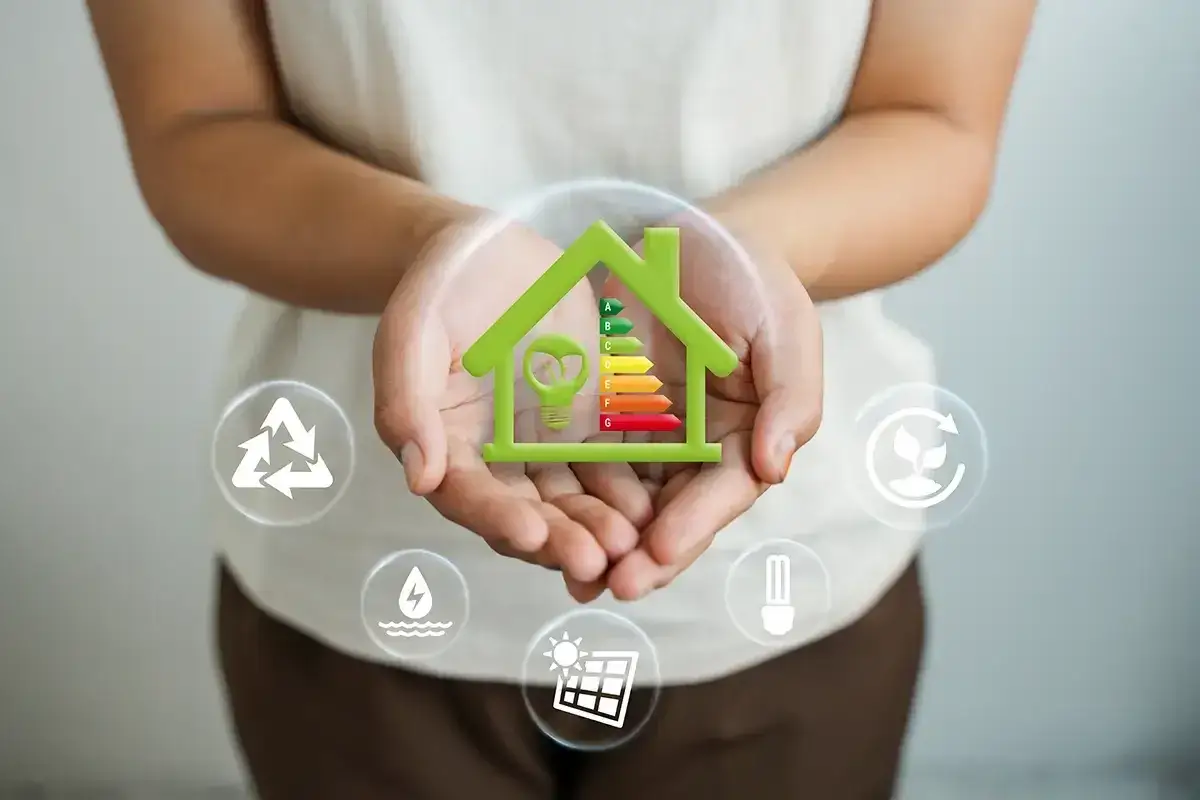 Zielony dom z ikonami symbolizującymi ekologię i efektywność energetyczną. Wewnątrz domu znajduje się żarówka i skala klasa energetyczna budynku (kWh/m²/rok).