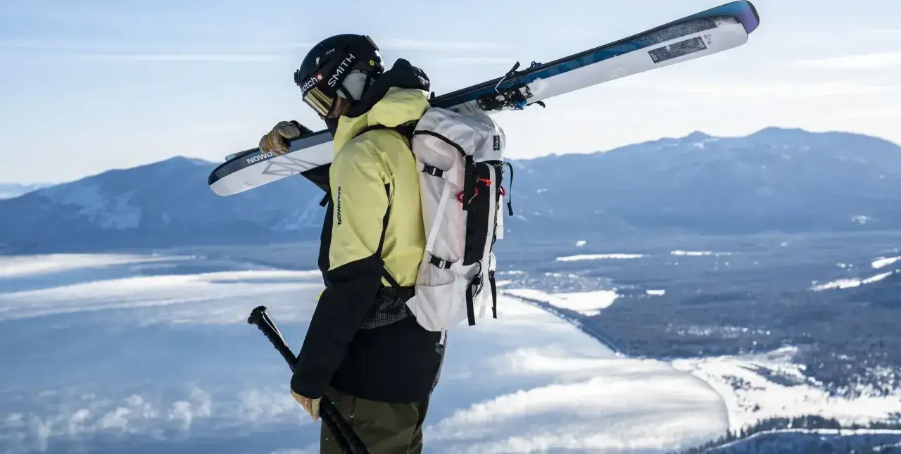 Narciarz w żółtej kurtce z nartami na ramieniu i plecakiem, na tle gór i jeziora.